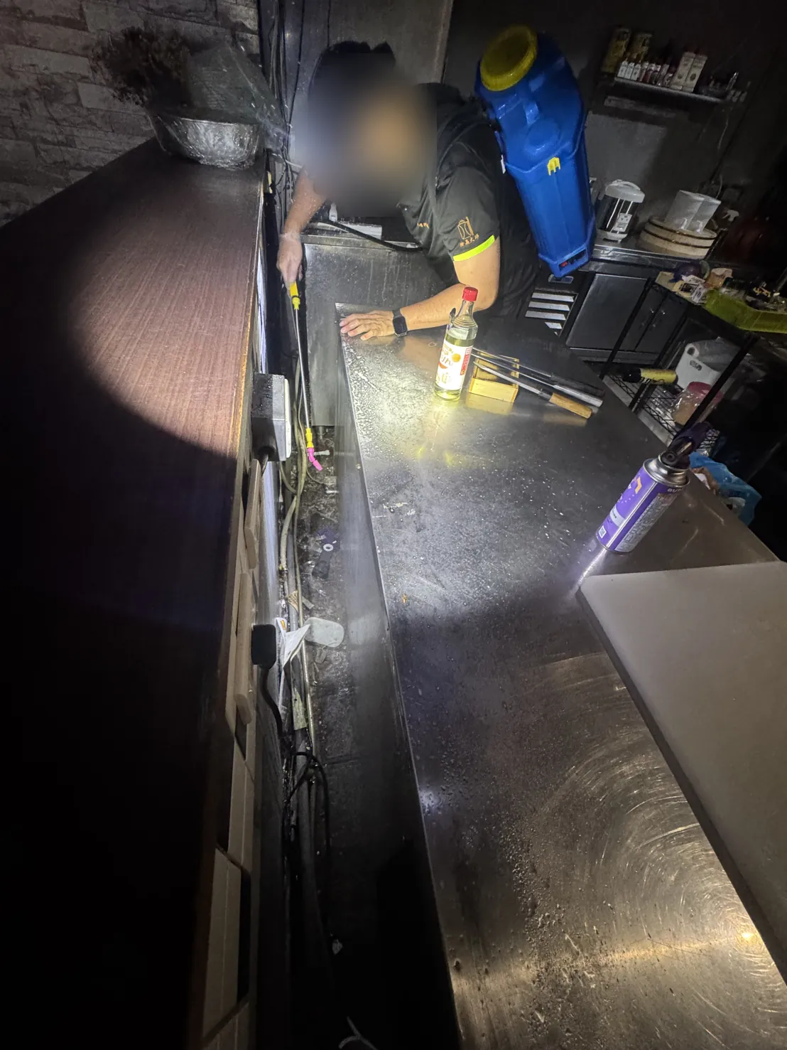 技師戴頭燈沿不鏽鋼工作檯與牆之間的縫隙檢查施藥（臉部已模糊）